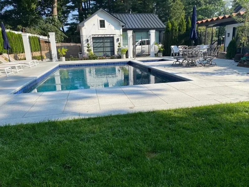 Pool with stone coping beside a lawn, pool house and patio dining set